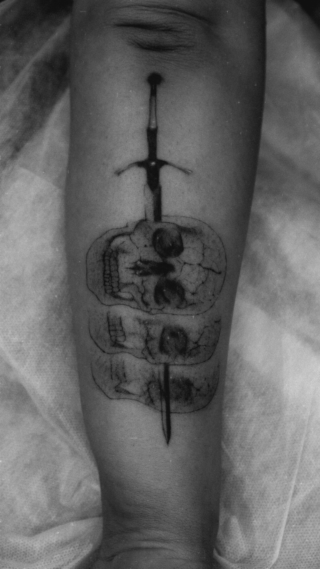 Black and grey forearm tattoo of a dagger extending through stacked skull-like forms.