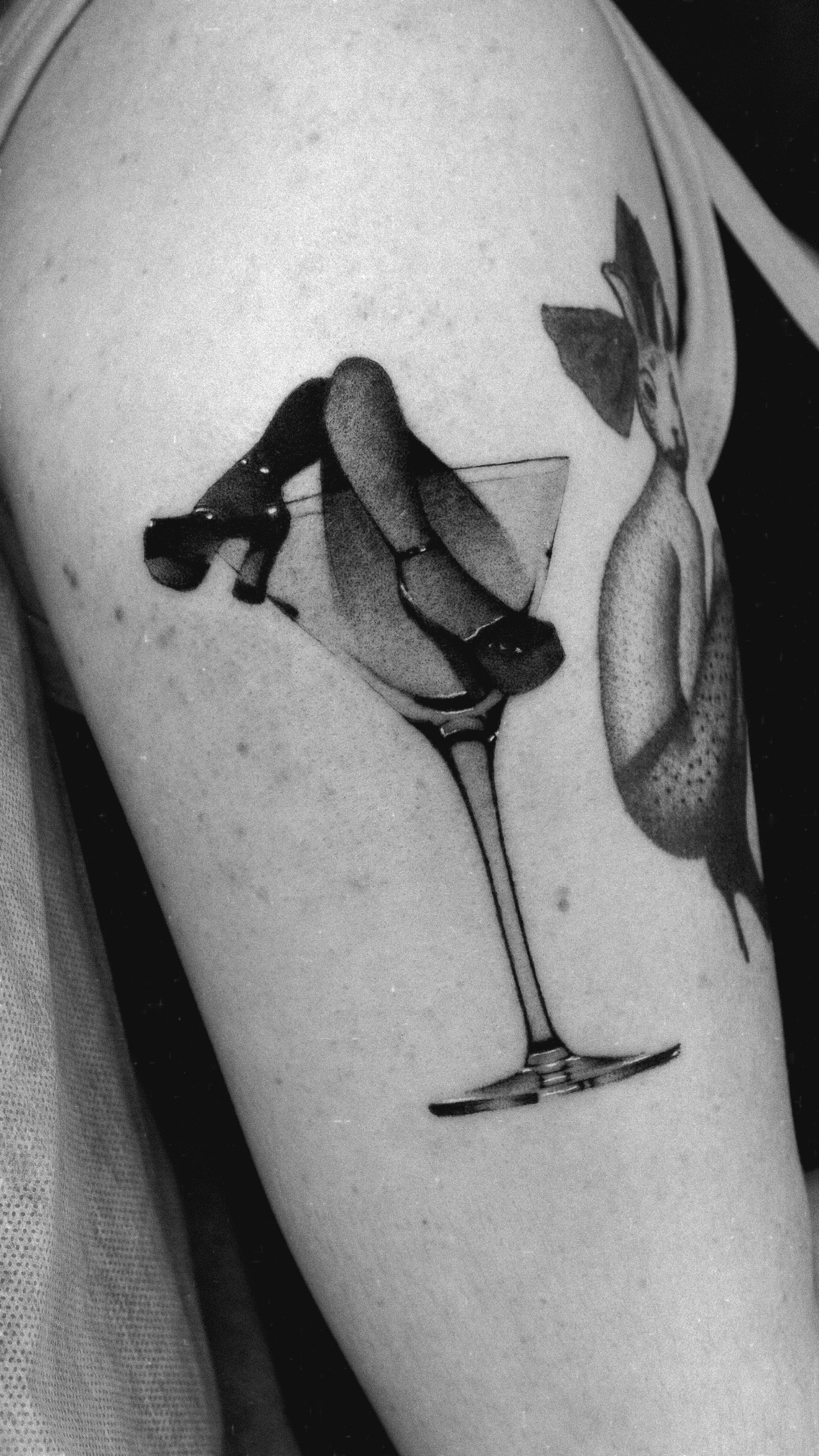 Black and grey arm tattoo of crossed legs rising from a stemmed glass.