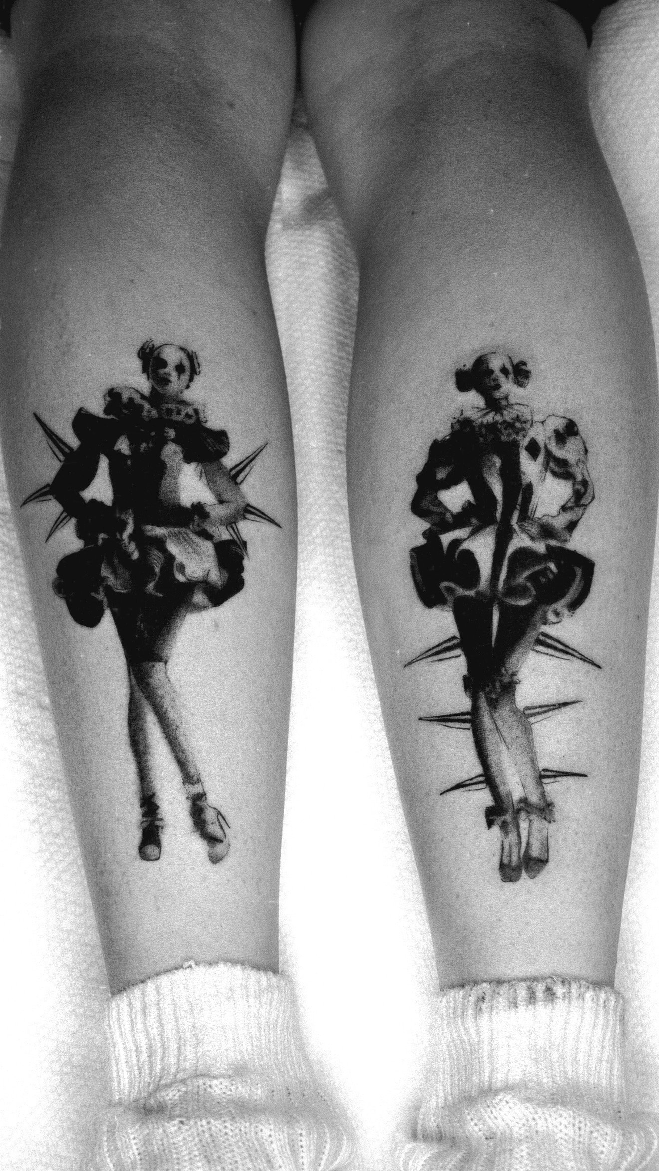 Paired black and grey lower-leg tattoos of elongated figures with dark floral and pointed shapes.